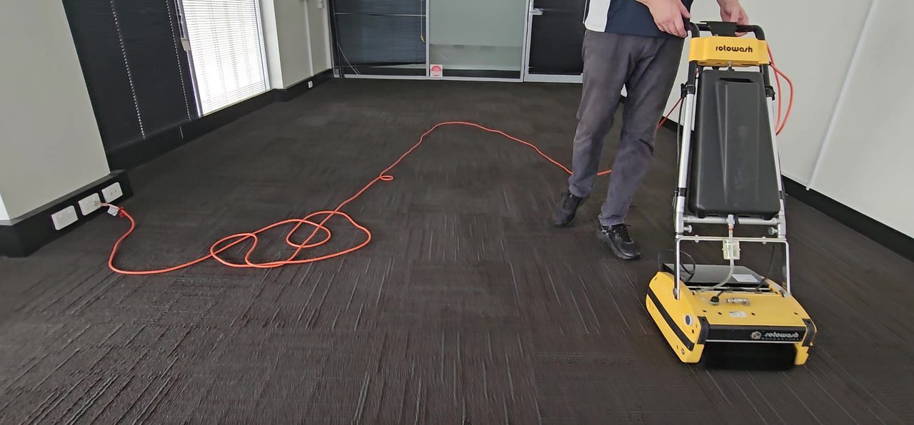A cylindrical brush machine scrubbing charcoal carpet tiles across an empty Perth office, its brushes working deep into the pile