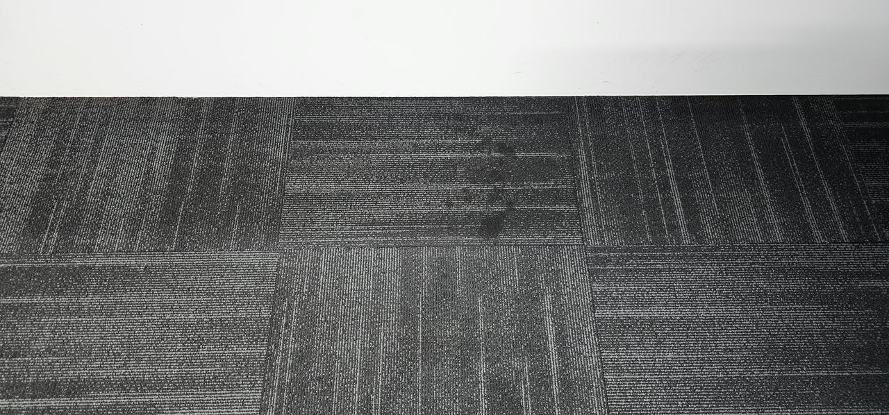 A dark greasy stain across two charcoal carpet tiles in a Perth office, the mark to clear before an audit, inspection or big event