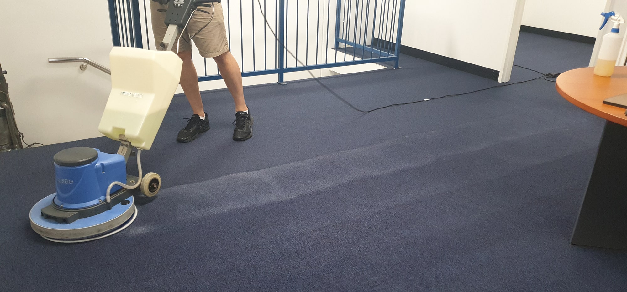 A scrubbing machine leaving a clean, bright pass across navy carpet near a Perth office stairwell