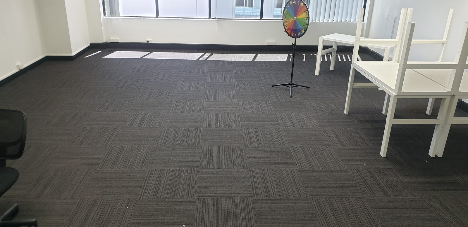 A cleared office room floored in dark grey carpet tiles, white desks stacked to one side and a rainbow-coloured wheel standing on a black tripod