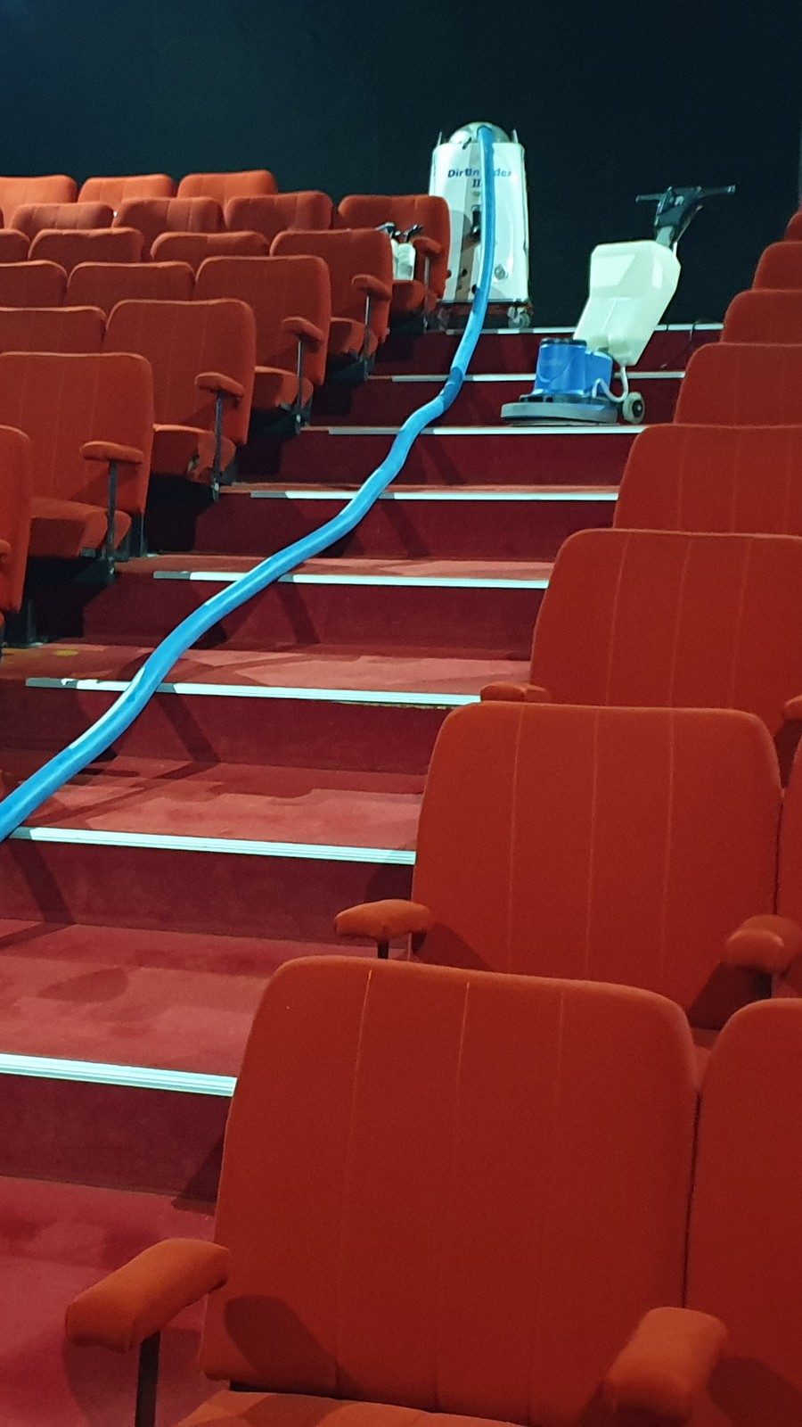 Steam cleaning hoses run up the aisle stairs of a Perth cinema between rows of red seats, the extraction unit at the top.
