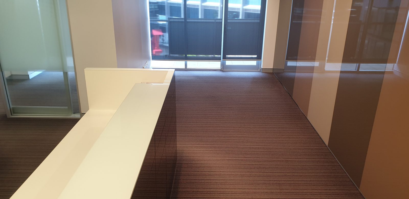 A glazed partition on the left shows grey carpet beyond it, dark brown ribbed carpet running past a gloss cream counter to a wall of glass