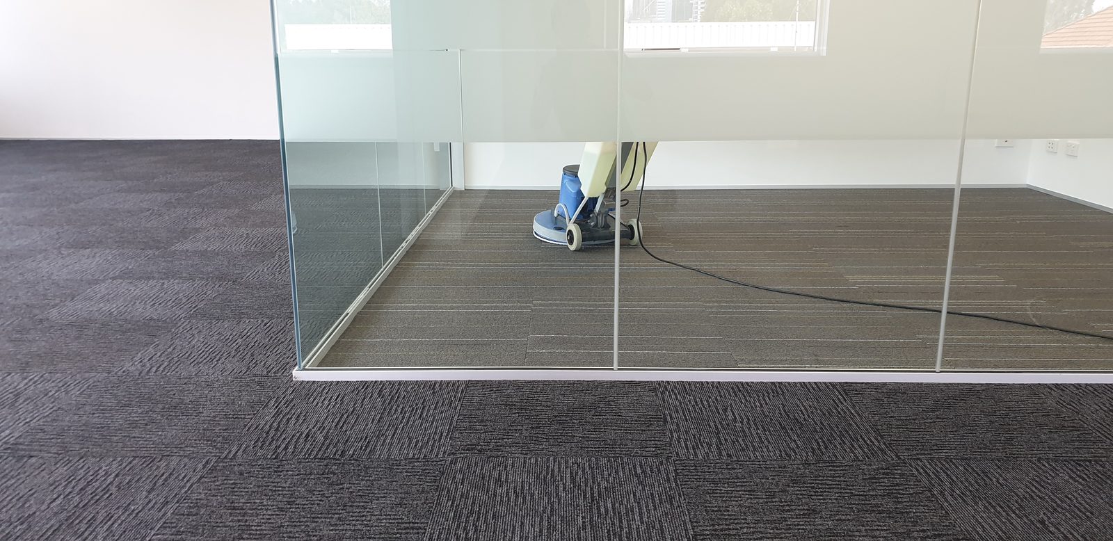 A floor machine with a blue drum stands beyond a glass partition, its lead trailing over grey-brown tiles while charcoal tiles fill the near side