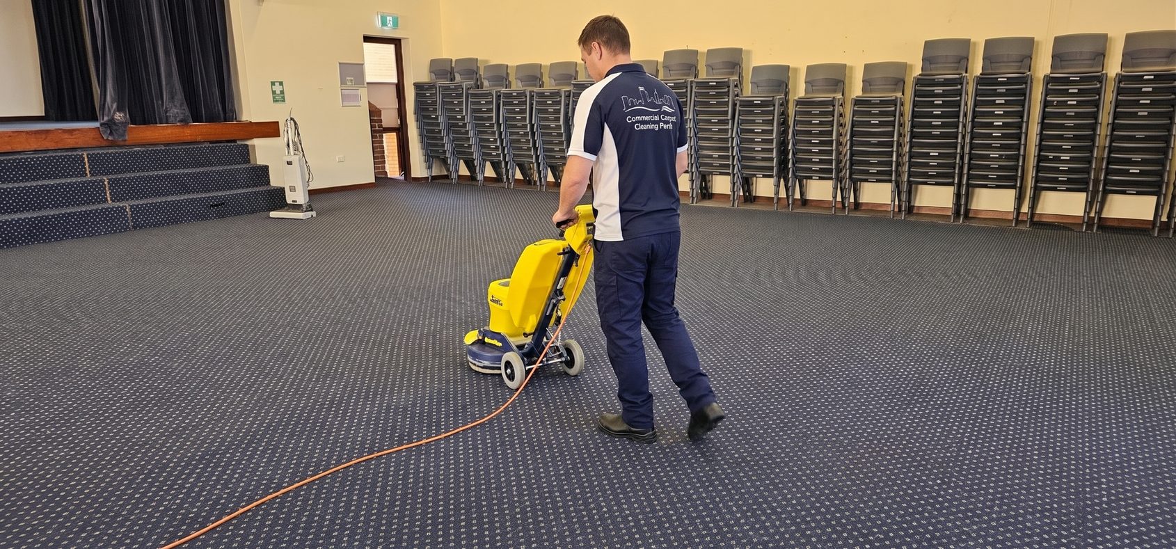 Our technician working a rotary machine across the carpet of a Perth function hall, the chairs stacked along the wall until the floor is handed back.