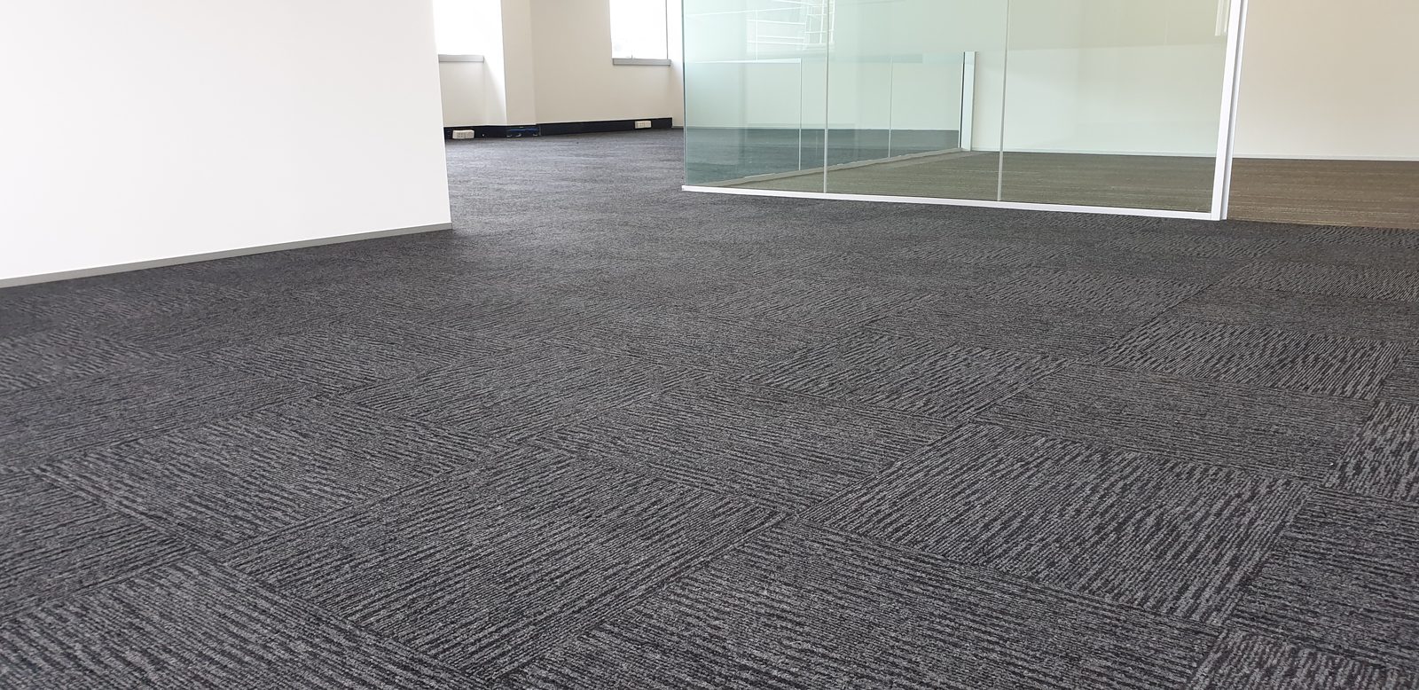 A white wall stops short at the left of a bare open-plan floor, dark grey carpet tiles running past it to glass panels set in a white track