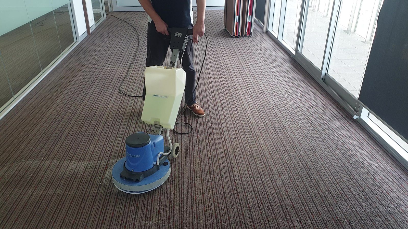 A wide office floor of maroon and grey pinstriped carpet running away between glazed walls, a technician behind the rotary machine holding a loop of its lead