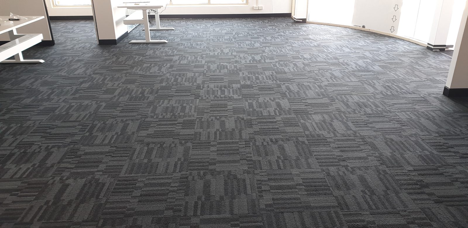 Carpet tiles laid in a quarter-turn grid so the grey pattern runs one way then the next, an empty office floor lit from the far windows